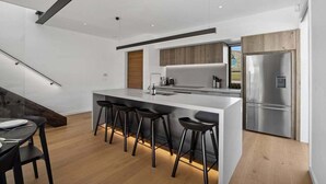 A modern kitchen with a central island, bar stools, a stainless steel refrigerator, and a wooden accent wall. - The Pisa (Cardrona)