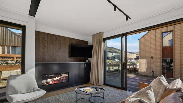 A modern living room with a fireplace, a flat-screen TV, and a large sliding glass door leading to an outdoor area. - The Pisa (Cardrona)