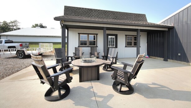 An outdoor patio area with a round table and four Adirondack chairs, a white building with a dark roof, and a truck parked nearby.