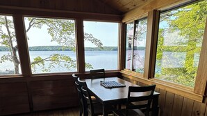A wooden cabin with a dining table and chairs, large windows with a lake view, and a cozy interior.