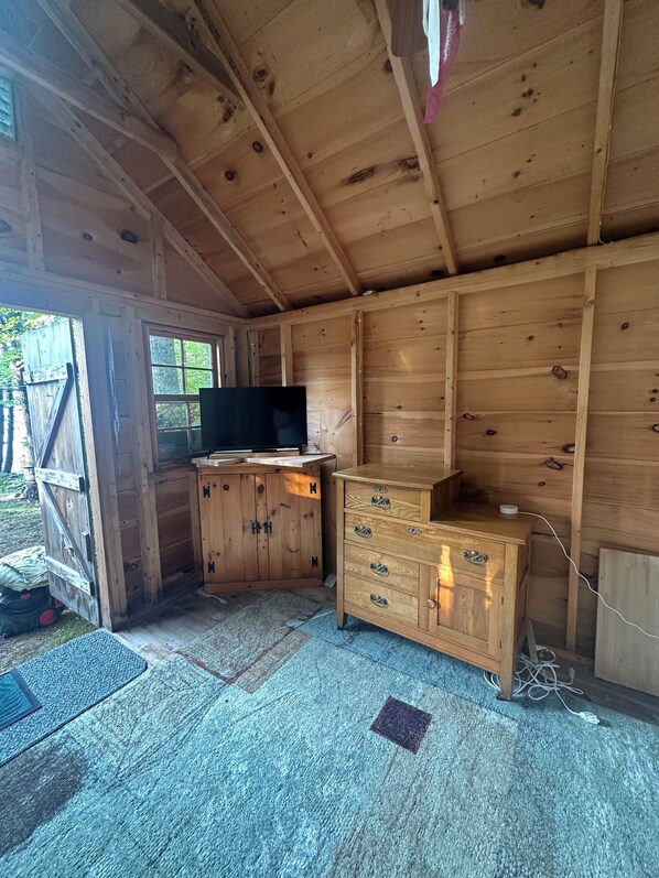 Interior de una cabaña de madera con una televisión sobre un mueble de madera, un escritorio pequeño y una ventana.