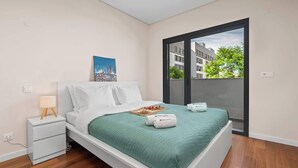 Une chambre à coucher moderne avec un lit, une table de chevet, une fenêtre donnant sur des immeubles et un tableau accroché au mur.