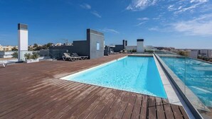 Une piscine sur le toit, dotée d’une terrasse en bois, de chaises longues et offrant une vue dégagée sur le panorama urbain.