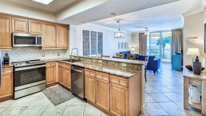 A modern kitchen with granite countertops, wooden cabinets, a built-in microwave, and a dishwasher. There is a dining area with blue furniture and a view of the outdoors. - 3BR ~ Beachfront ~ Huge Balcony ~ 2 Pools ~ Luxury (Fort Walton Beach)
