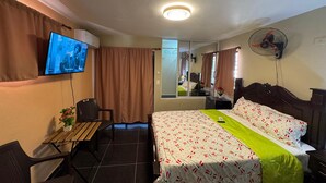 Una camera d'albergo con un letto, una scrivania con sedia, una televisione, uno specchio, una ventola e una finestra con tende.