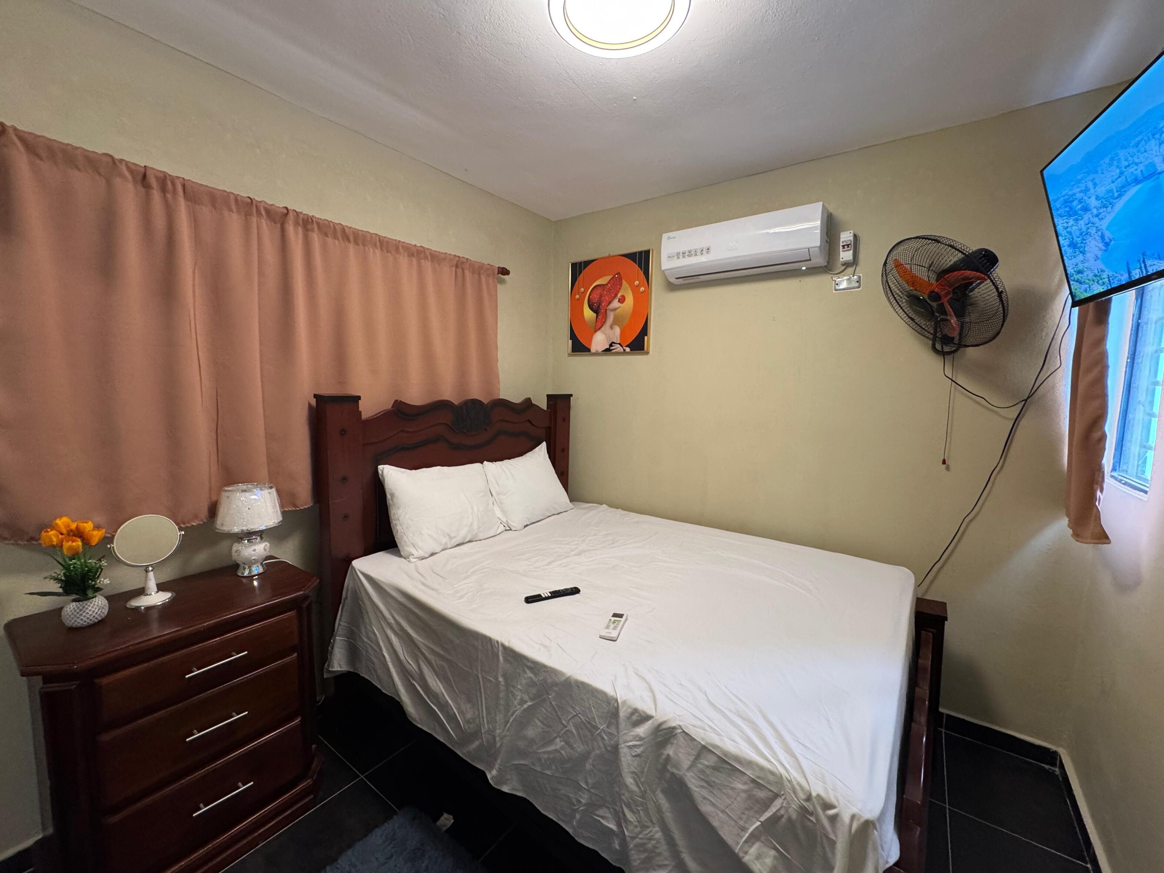 Habitación de hotel con cama, mesita de noche con lámpara, espejo, cómoda, aire acondicionado, ventilador de pared y televisor en la pared.