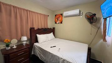 Habitación de hotel con cama, mesita de noche con lámpara, espejo, cómoda, aire acondicionado, ventilador de pared y televisor en la pared.