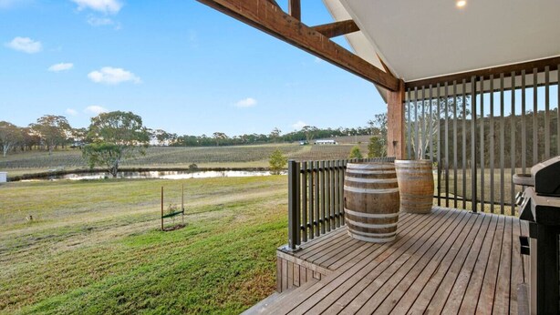 Eine Holzterrasse mit Grill und zwei Fässern, mit Blick auf ein Wiesenfeld und ein Gewässer.