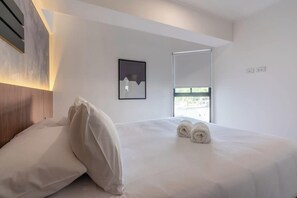 Una habitación de hotel moderna con una cama, un ventanal grande, un espejo montado en la pared y un cuadro enmarcado.