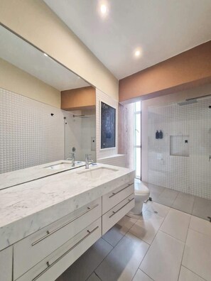Uma casa de banho moderna com um espelho grande, duas lavatórios, chuveiro e sanita.