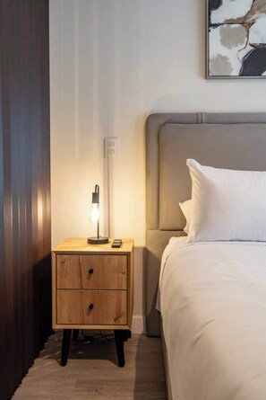 Ein ordentlich bezogenes Bett mit weißer Bettwäsche, eine Nachttischlampe, ein Nachttisch mit Schubladen und ein Wandbild.