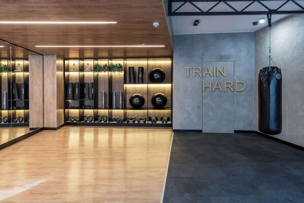 Un gimnasio con piso de madera, un saco de boxeo y una pared con la inscripción 'TRAIN HARD'.