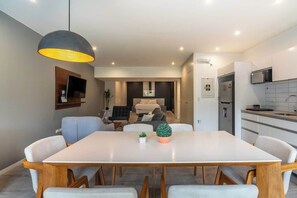 A modern kitchen with a microwave, a dining area with a white table and chairs, and a living area with a sofa and a TV.