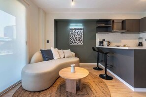 Un espace de vie moderne comprenant un canapé incurvé, une table basse ronde, un coin cuisine avec un bar et un élément décoratif mural.