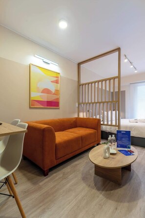 A hotel room with a sofa, a round coffee table, a bed with white linens, a wall-mounted artwork, and a wooden partition.