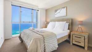 A bedroom with a large window, a bed with white linens, a wooden nightstand, and a lamp.