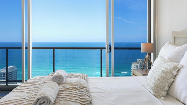 A hotel room with a large balcony, a bed with white linens, a lamp, and a view of the ocean.