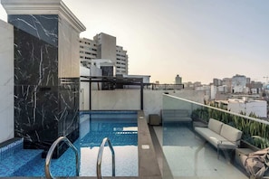 Una piscina en la azotea con barandilla de vidrio, una zona de estar con cojines y vistas a la ciudad.