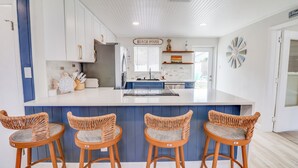 A kitchen with a central island, blue cabinets, wicker chairs, and a sign reading "BEACH HOUSE".