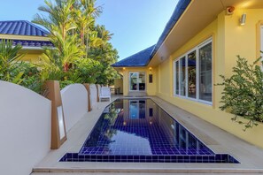 A swimming pool with a tiled deck, surrounded by a white wall and a yellow building with large windows and a blue roof.