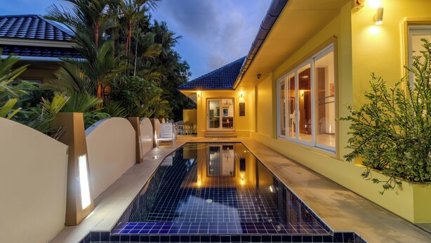 Une piscine avec une terrasse carrelée, un patio couvert et un bâtiment doté de grandes fenêtres et d’une porte.