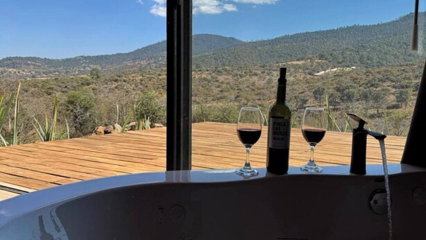 Banheira de hidromassagem com duas taças de vinho tinto e uma garrafa em deck de madeira, com vista para uma paisagem montanhosa.