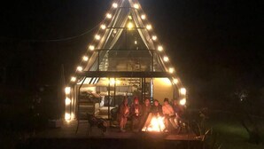 Uma estrutura triangular com luzes de cordão, uma lareira e pessoas sentadas ao redor.