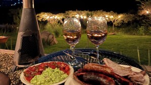 Uma mesa posta ao ar livre com uma churrasqueira, duas taças de vinho, um prato de salsichas e uma tigela de guacamole.
