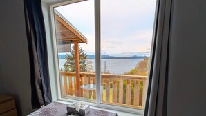 A room with a large window offering a view of a wooden deck and a body of water. - The Founder's Vista Suite Spacious, Panoramic Ocean Views (Alert Bay)