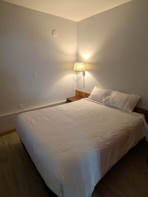 A neatly made bed with white linens in a simple room with a single bedside lamp. - The Sitka Suite | Central Apartment, Waterfront, 2 Beds (Alert Bay)