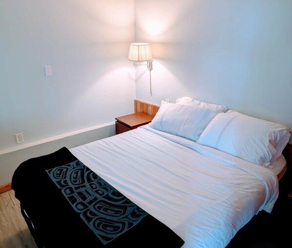 A neatly made bed with white linens and a black throw blanket, a bedside lamp, and a wooden nightstand. - The Sitka Suite | Central Apartment, Waterfront, 2 Beds (Alert Bay)