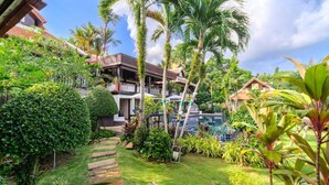 A lush garden with a pool, palm trees, and a building with a balcony.