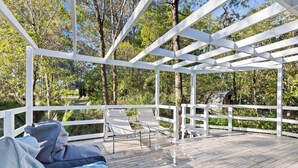A covered outdoor deck with a white railing, a lounge chair, and a small table. - A Classic Beach House - Waterfront Access and Short Walk to Beach (Mossy Point)