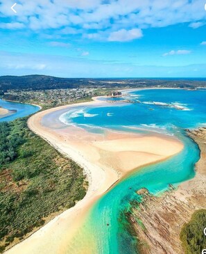 Vue aérienne d’une zone côtière avec une plage de sable en forme de courbe, des eaux turquoise et limpides, et une ville située au loin.