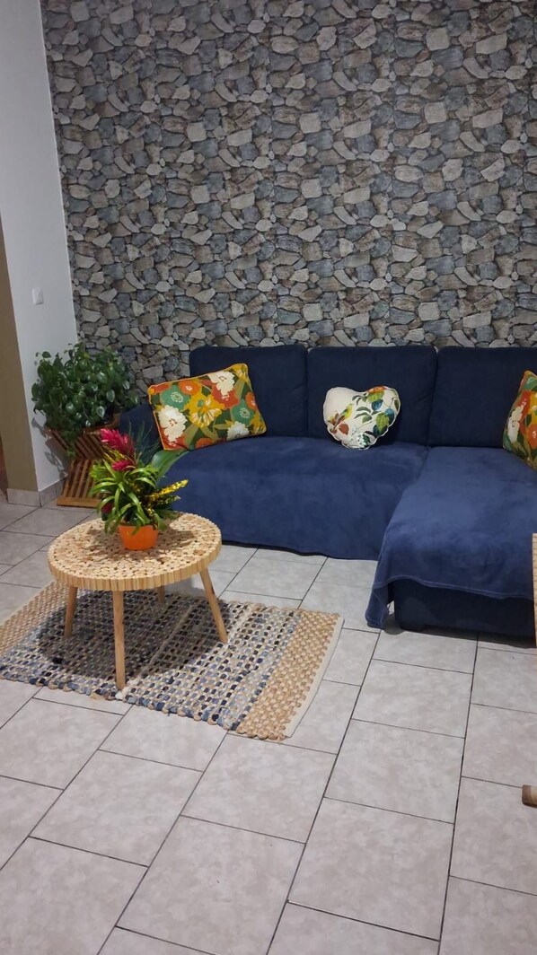 A living room with a stone wall, a blue sofa, colorful cushions, a round wooden table with a plant, and a patterned rug.