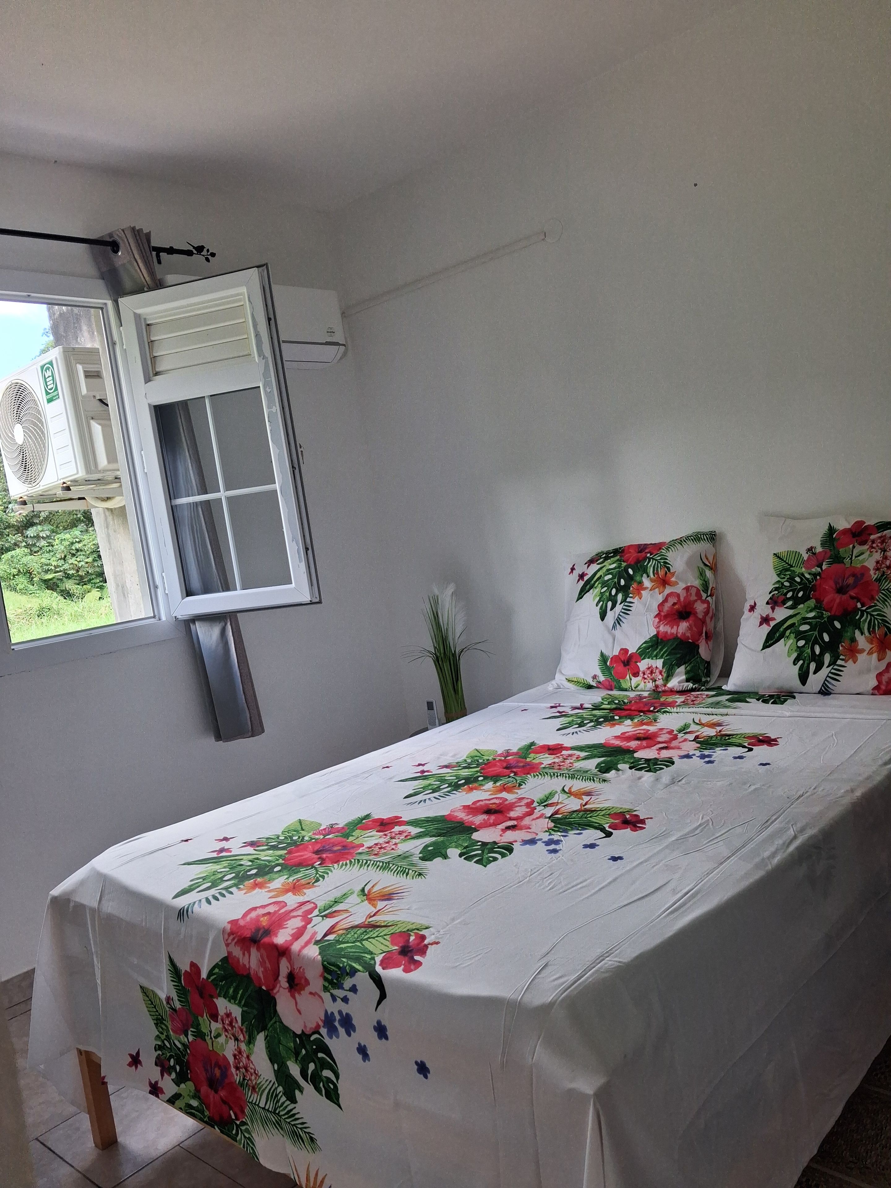 A bedroom with a bed, floral bedspread, pillows, a window with curtains, and an air conditioning unit.