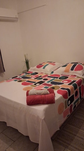 A bed with a white bedspread and colorful pillows, a small plant on a side table, and a wall-mounted air conditioner.