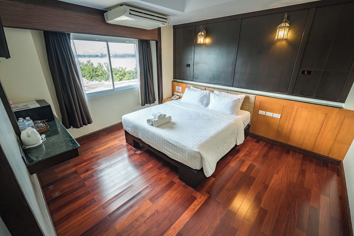 A hotel room with a large bed, wooden flooring, a window with curtains, a bedside table with a lamp, and a view of the outdoors.