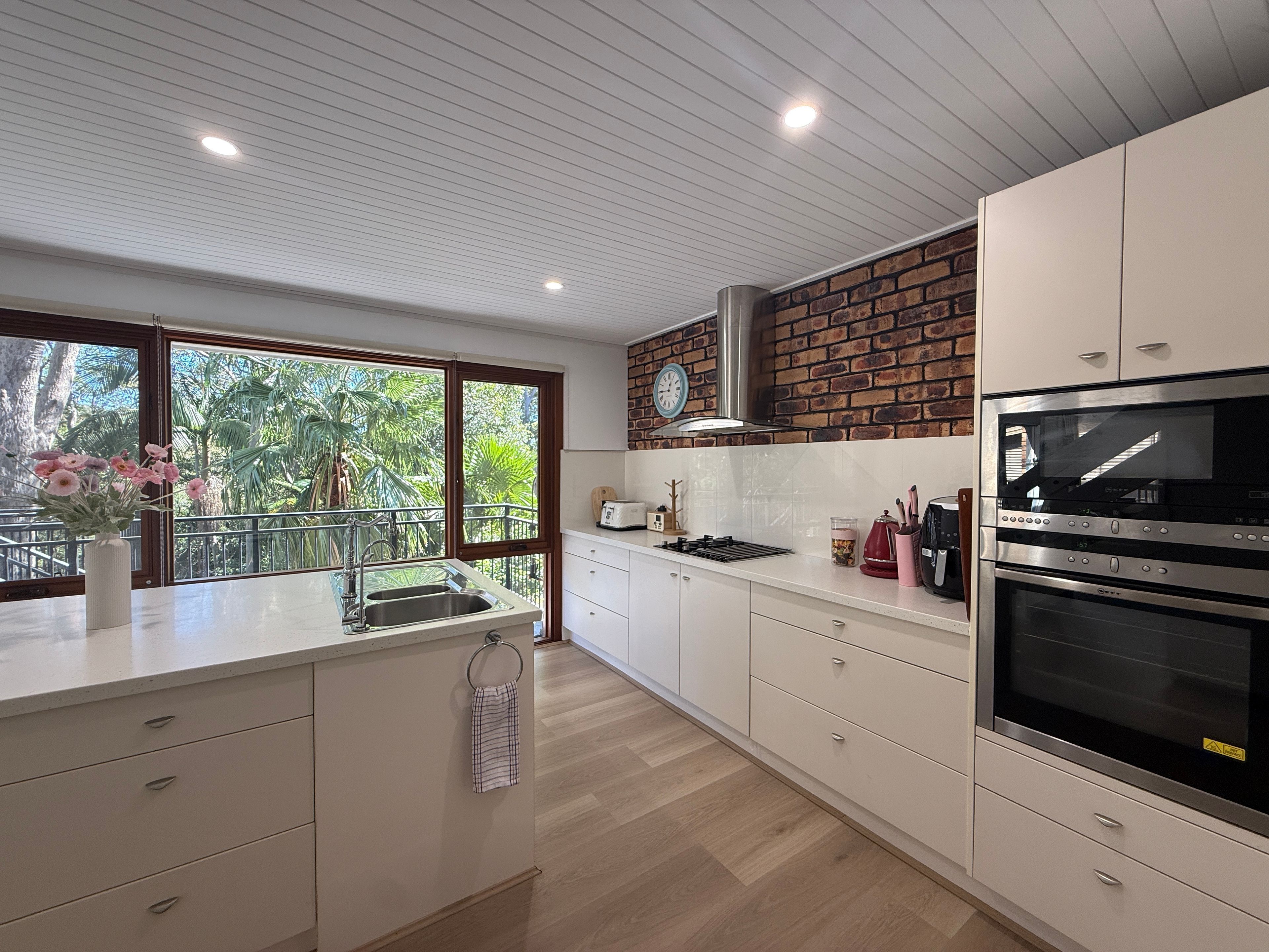A modern kitchen with a large island, a brick accent wall, and a view of greenery through the windows.