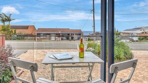 Une terrasse avec deux chaises blanches, une table, une bouteille de vin et un verre. En arrière-plan, on aperçoit des maisons aux toits rouges et un ciel dégagé.