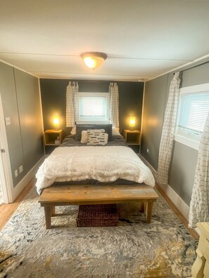 A bedroom with a large bed, bedside lamps, a wooden bench, and a window with curtains. - The Sweetheart Cabin.  A Charming 1-bedroom cabin with a super comfy bed! (Livermore)