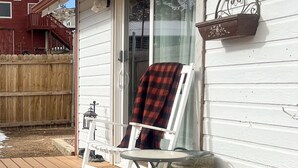 A wooden deck with a folding chair and a small table, a white house with a red barn in the background, and a clear sky.