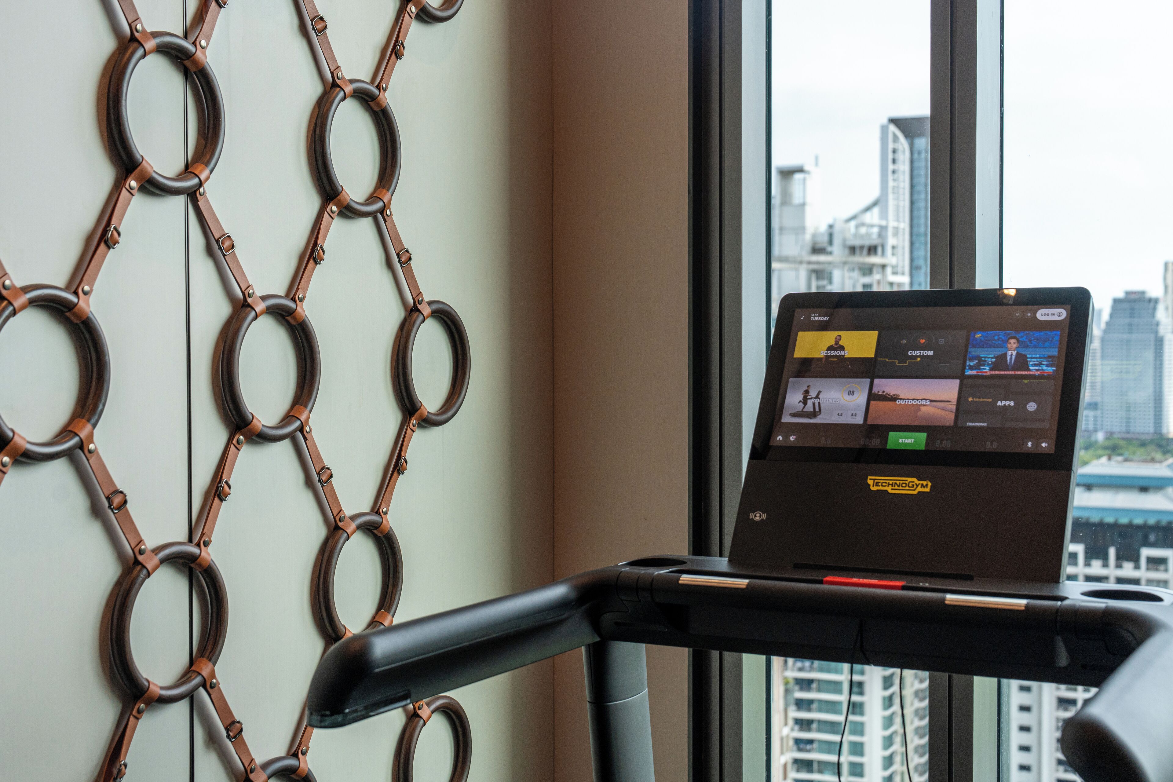 Ein Laufband mit einem Laptop, der Trainingsmöglichkeiten anzeigt, eine Maschendrahtwand und ein Blick auf die Stadt durch ein Fenster.