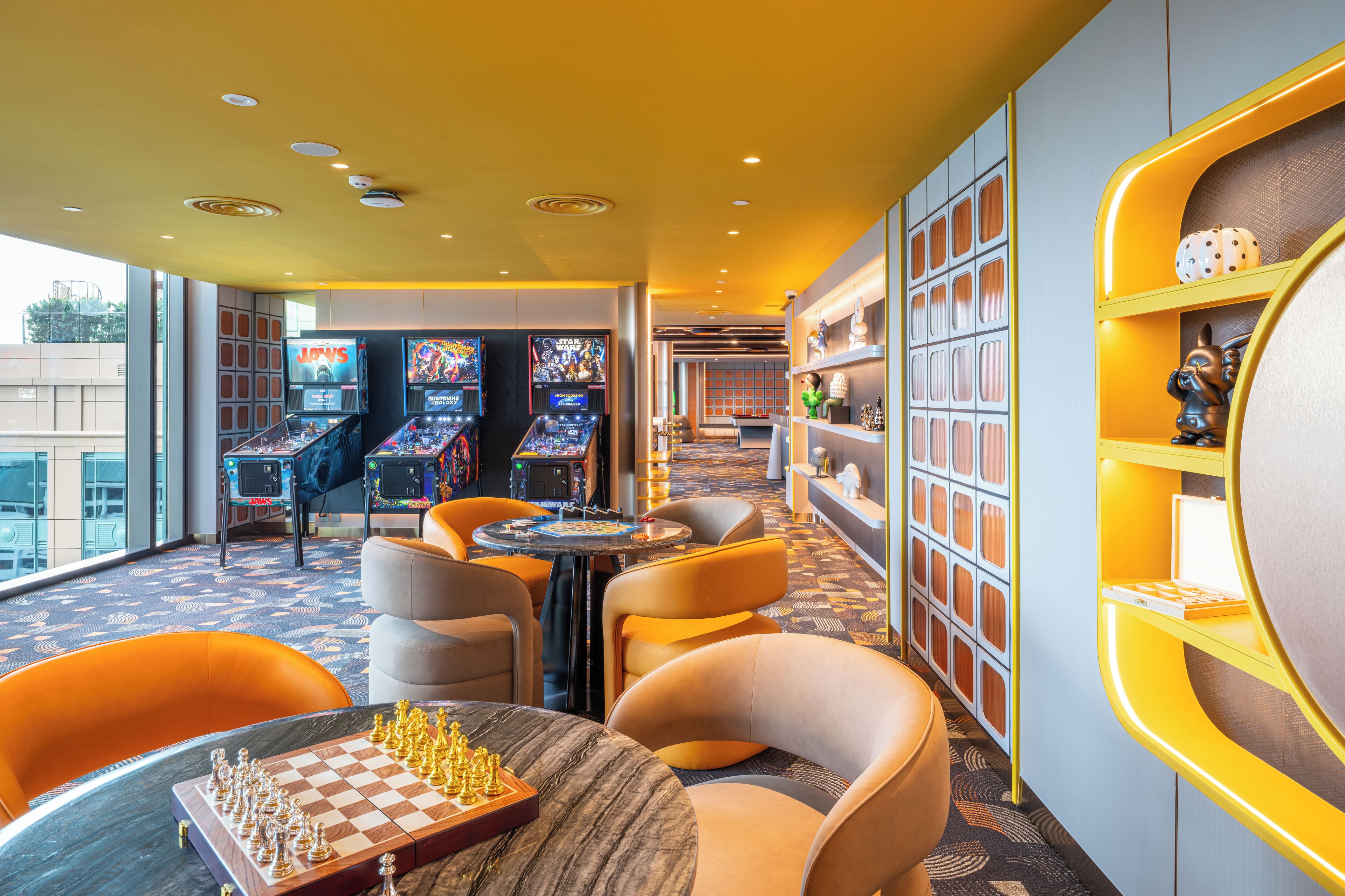 Un área de lounge moderna con un gran tablero de ajedrez, varias máquinas de arcade, asientos cómodos y un ventanal con vistas a la ciudad.