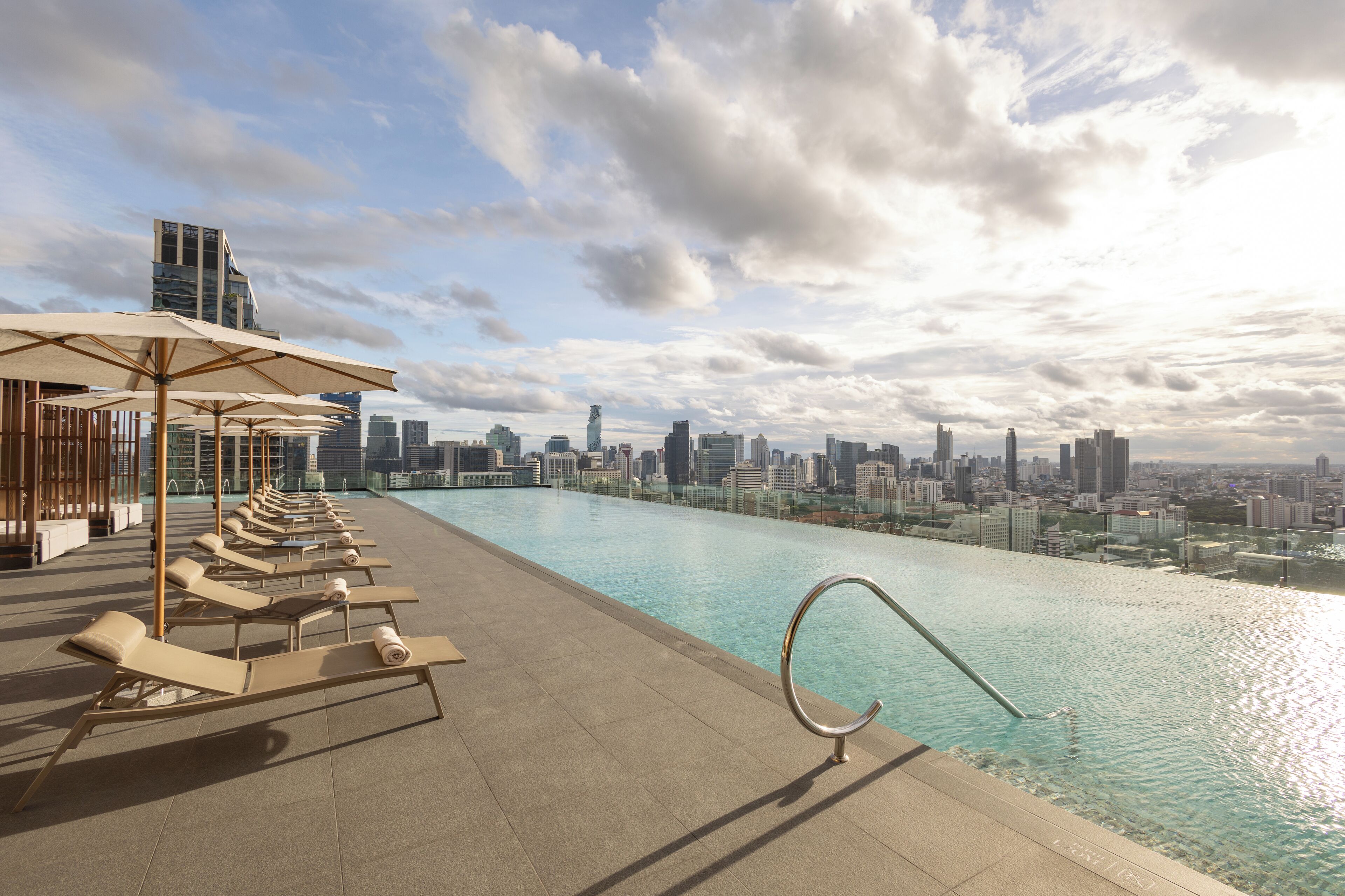 A rooftop pool with lounge chairs and umbrellas, overlooking a city skyline.