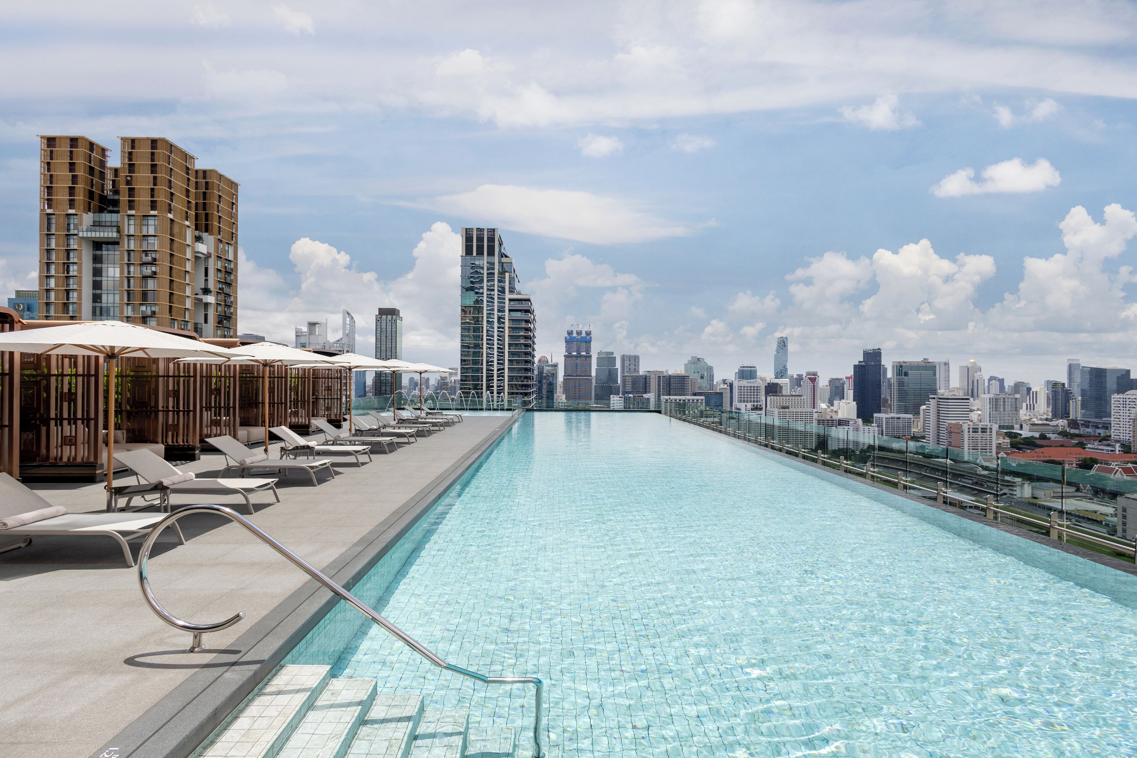 A rooftop infinity pool with lounge chairs and umbrellas, overlooking a city skyline.