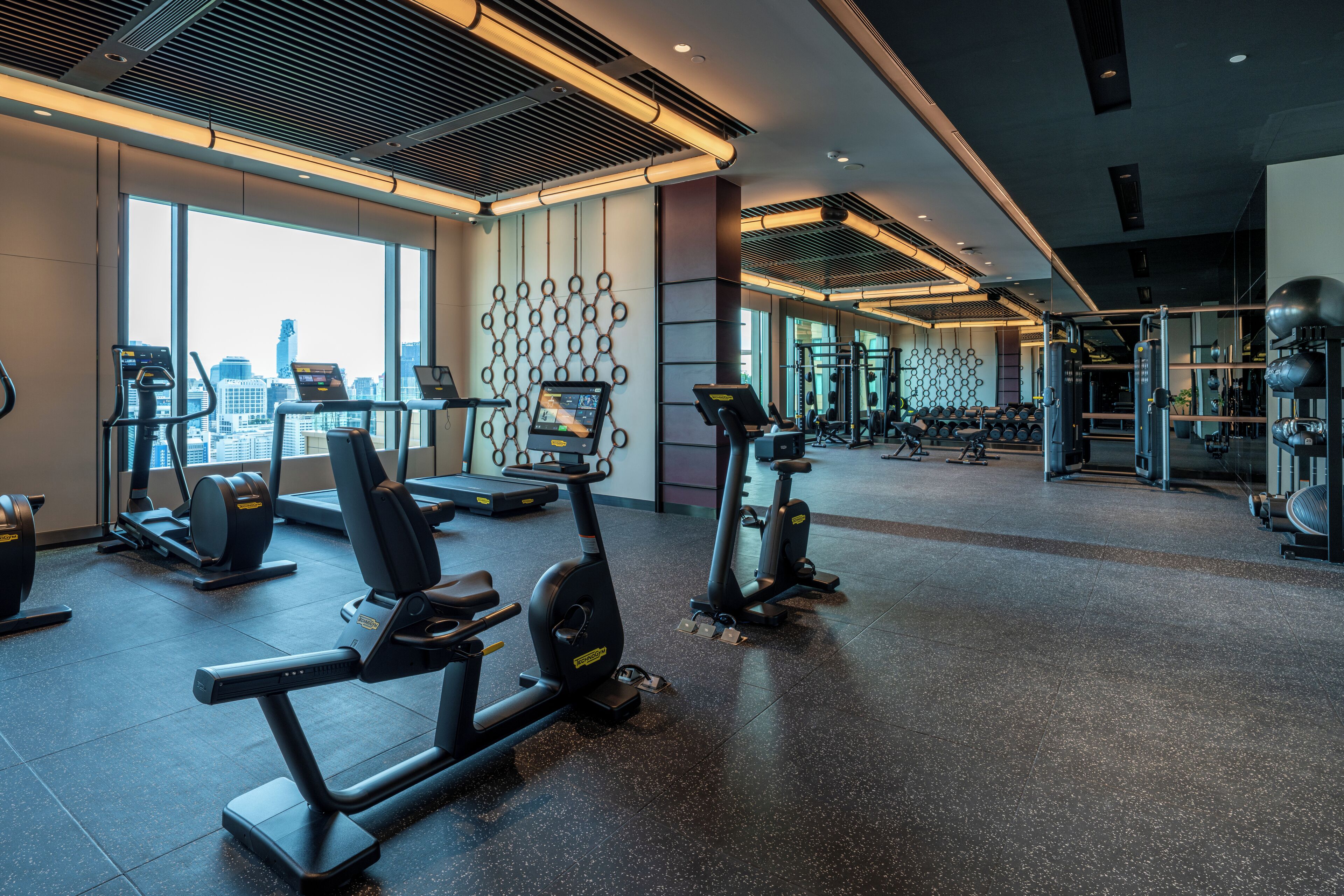 Un gimnasio moderno con máquinas de cardio, equipo de levantamiento de pesas y vistas a la ciudad a través de amplios ventanales.