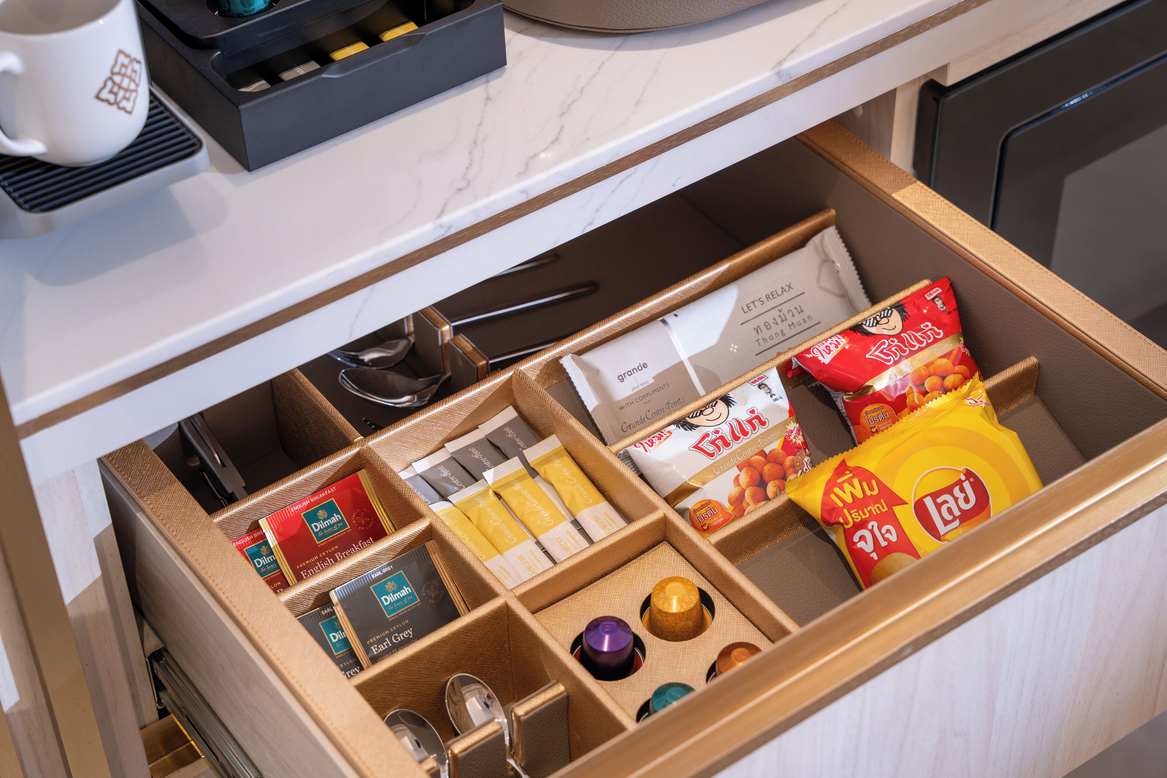 A drawer with compartments containing various items such as a white mug, a box of tea bags, a packet of snacks, and a small container.