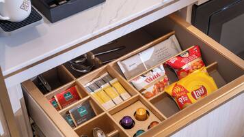 A drawer with compartments containing various items such as a white mug, a box of tea bags, a packet of snacks, and a small container.