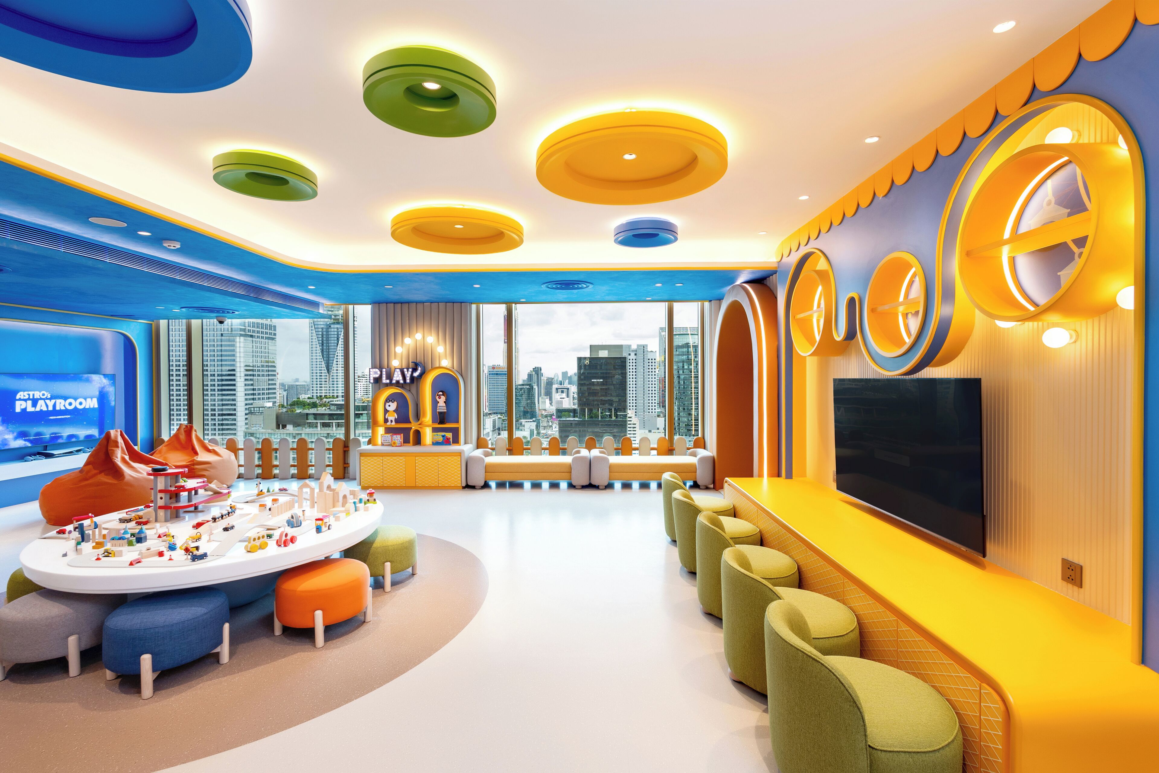A modern playroom with a city view, colorful ceiling lights, and a large TV screen.